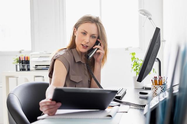 Une femme travaillant à son bureau, regardant une tablette tout en passant un appel téléphonique